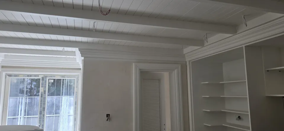 Beyaz kirişli lambri tavan, panjurlu kapı ve gömme açık raflarla villa odası